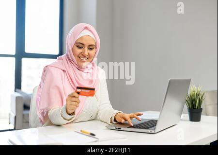 Young Muslim Arab mixed-race businesswoman in hijab sitting at the desk and entering typing credit card number on laptop, feeling excited to purchase items from an online store, order food, pay bills Stock Photo