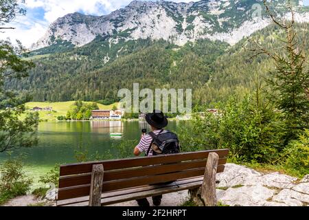 Hiker sits on a bench and films the landscape with the small camera, Hintersee, Ramsau, in the back Reiteralpe, Berchtesgaden, Berchtesgaden Alps, Berchtesgaden National Park, Berchtesgadener Land, Upper Bavaria, Bavaria, Germany, Europe Stock Photo