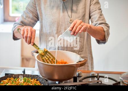 Hands of a woman on a gas stove pours coconut milk from a jug into a pot of boiled sweet potatoes Stock Photo