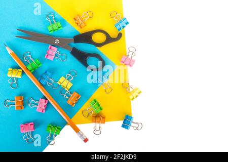 Stationary flat lay on blue and yellow paper with white background and empty space on one side. Lots of small colored paper clips, little scissors, an Stock Photo