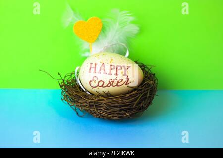 Easter egg with a Happy Easter message in a bird's nest on a green and blue background. Gold decorative heart. Festive Easter background. High quality Stock Photo