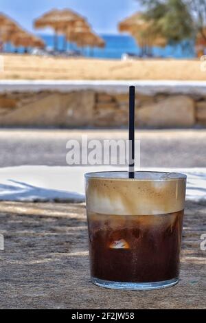 Closeup of an iced coffee with a straw in front of the mylopotas beach in Ios Greece Stock Photo