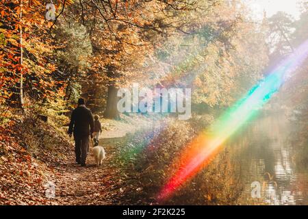 Man, child and dog walking on canal towpath with rainbow flare in fall Stock Photo
