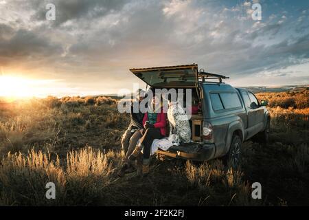 Cheerful couple sitting with dog at trunk of off-road vehicle on field during vacation Stock Photo