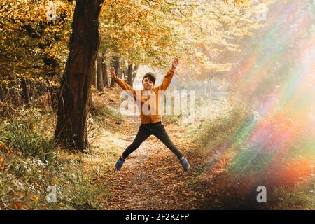 Boy leaping in the air on path under tree with rainbow light flare Stock Photo