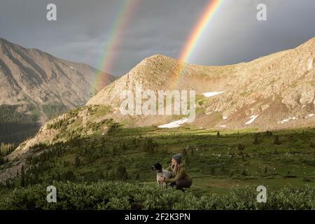 Woman stroking dog while double rainbow and mountains in background Stock Photo