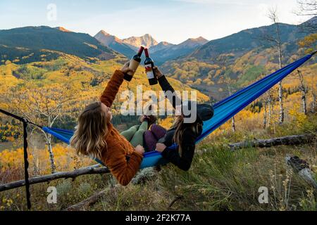 Female friends toasting wine bottles while relaxing on hammock in forest Stock Photo
