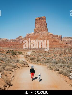 Rear view of woman walking with dog on dirt road leading towards sandstone formations against clear sky Stock Photo