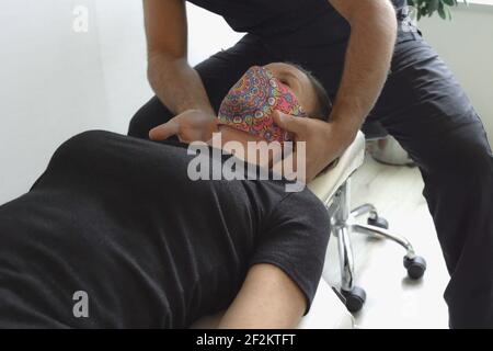 Anonymous osteopath sitting on chair massaging neck of female patient with face mask lying down on stretcher. Physiotheraphy session Stock Photo