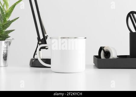 Enamel mug mockup with a lamp, aloe vera plant and workspace accessories on a white table. Stock Photo