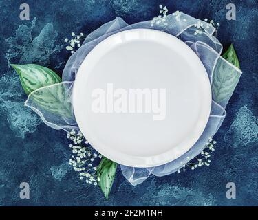 Spring season composition with a white plate and flowers on a blue background. Romantic, floral table setting with large leaves and gypsophila. Weddin Stock Photo