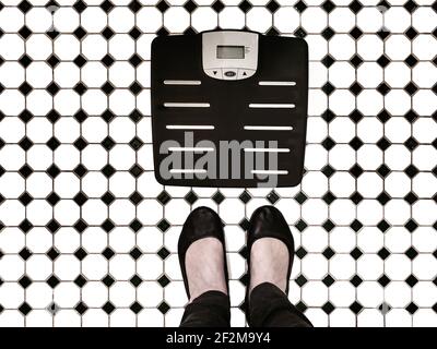 Body weight scales on tiled bathroom floor with woman's feet beside it like shes going to step on them - top view Stock Photo