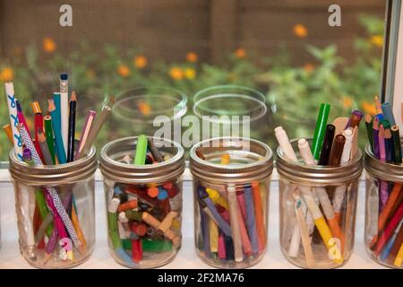 Canning jars filled with crayons, pastels and markers sitting on an open windowsill with flowers outside seen through a window screen Stock Photo