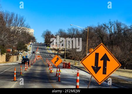 A city road by a park under construction with four lanes reduced to two with in focus signs in foreground and blurred buildings and trees in distance Stock Photo