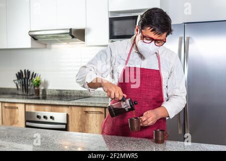 Man in mask and red apron pouring coffee from a French press into a cup in a home kitchen or cafeteria. Stock Photo