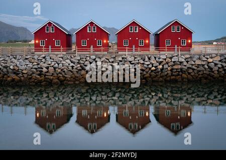 classic red fishing cottages in perfect symmetry sitting along a stone jetty with a slightly blurred reflection. Classic red houses of northern norway Stock Photo
