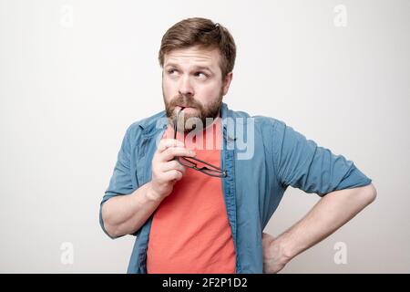 Pensive man holds glasses in mouth and tries to remember something, on a white background. Stock Photo