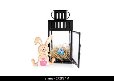 Wooden Easter bunny with a colorful Easter egg in a nest within a decorative lantern, isolated on white background. Easter celebration concept. Stock Photo
