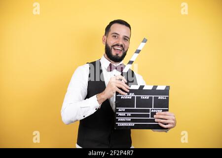 Young man with beard wearing bow tie and vest holding clapperboard very happy having fun Stock Photo