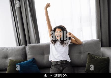 Satisfied beautiful African American girl sits on the sofa in living room. Happy young woman chilling and smiling at home Stock Photo