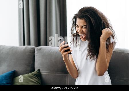 Overjoyed young woman use smartphone, receive a good message. Happy african american girl get a promotion on work or passing exams. Joyful female freelancer sit on the sofa, enjoy good results Stock Photo