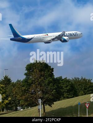 London, United Kingdom, July 2019 - Kuwait Airlines, Boeing 777 taking off into a blue sky with broken clouds with trees and grass in the foreground. Stock Photo