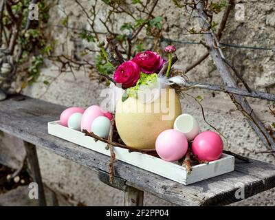 Easter eggs from hens, geese and ostrich in pastel colors on an elongated white wooden tray with a bouquet of ranunculus in the ostrich egg, on a raw wooden bench in front of the farmhouse wall Stock Photo