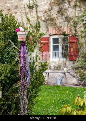 Easter decoration with ranunculus and Easter egg in a terracotta flower pot on a wooden post decorated with cut fruit branches and colored ribbons in front of a farmhouse Stock Photo