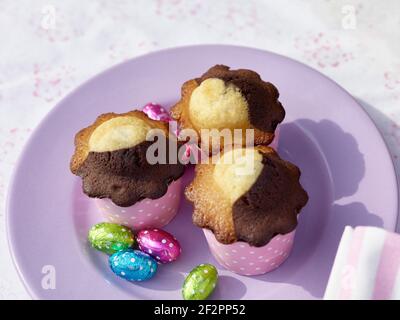 Three muffins on a lilac-colored plate with four chocolate Easter eggs wrapped in foil Stock Photo