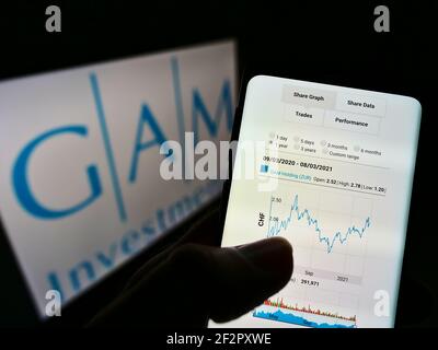 Person holding mobile phone with website and stock chart of Swiss company GAM Investments on screen with logo. Focus on center of phone display. Stock Photo