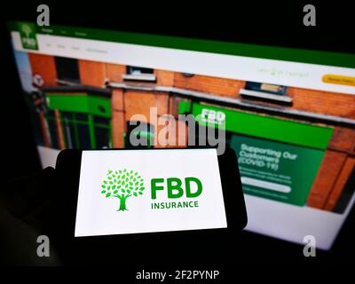 Person holding smartphone with logo of Irish insurance company FBD Holdings plc on screen in front of website. Focus on phone display. Stock Photo