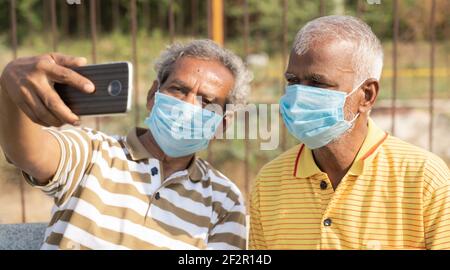 elderly friends with medical face mask taking selfie while sitting at park - concept of happy senior people friendship and meetup after lockdown due Stock Photo