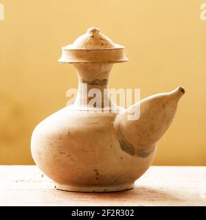 An ancient clay jug isolated on wooden table on blurred cream background. Ancient clay jug is display in the local museum of Sing Buri, Thailand. Stock Photo