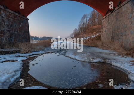 an old stone bridge box and reflection in the water just below the bridge on an uneven dirt road that is clear with ice and melting snow forming water Stock Photo