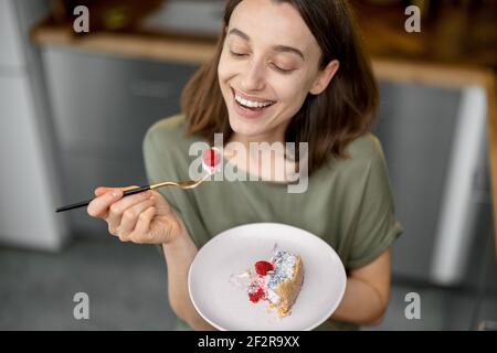 Young woman enjoys a sweet dessert, having a snack on a kitchen at home. Home made recipe of cheesecake with raspberry. Healthy cake. Stock Photo
