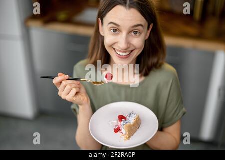 Young woman enjoys a sweet dessert, having a snack on a kitchen at home. Home made recipe of cheesecake with raspberry. Healthy cake. Looking and smiling at camera. Top view. Stock Photo