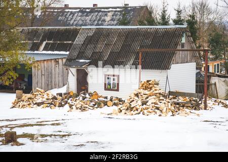 Pile and wall of cut birch wood and stacked wood logs ready for winter, standing on the snow near a Lithuanian typical wooden house in winter Stock Photo