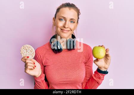 Beautiful caucasian sports woman holding green apple and rice crackers relaxed with serious expression on face. simple and natural looking at the came Stock Photo