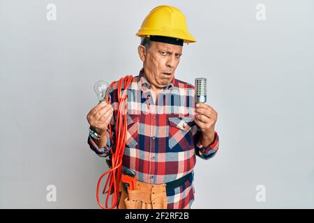 Senior hispanic man wearing handyman uniform holding led lightbulb and incandescent bulb in shock face, looking skeptical and sarcastic, surprised wit Stock Photo