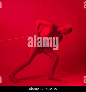 An anxious red man in a red leotard, dragging a heavy burden, pulling a heavy load with a rope. The concept of stress and burnout at work. Stock Photo