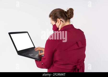 Rear photo of an overweight woman working online using a laptop laptop, wearing a protective textile mask. Working during a pandemic. Stock Photo