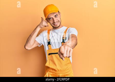 Hispanic young man wearing handyman uniform smiling doing talking on the telephone gesture and pointing to you. call me. Stock Photo