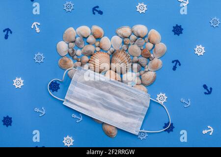 Heart-shaped shells with a medical mask on a blue background. Marine theme. top view Stock Photo