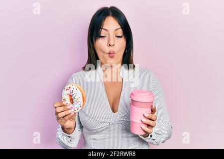 Young hispanic woman eating doughnut and drinking coffee making fish face with mouth and squinting eyes, crazy and comical. Stock Photo