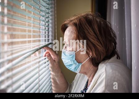 Sad lonely senior woman with face mask looking through window during covid-19 pandemic quarantine and lockdown Stock Photo