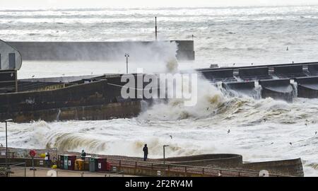 Brighton UK 13th March 2021 - People on the beach watch as waves crash over Brighton Marina as strong winds batter the south coast again today : Credit Simon Dack / Alamy Live News Stock Photo