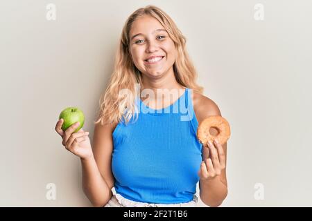 Young blonde girl holding green apple and donut smiling with a happy and cool smile on face. showing teeth. Stock Photo