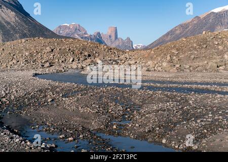 Wild river winds through remote, stony arctic landscape. A big moraine and iconic mountains on the horizon. Mt. Asgard, Akshayuk Pass, Nunavut, Canada Stock Photo