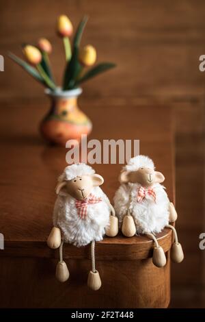 Funny sheep toys on wooden table, vase with tulips in background. Indoors decoration for Easter holidays Stock Photo