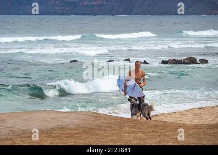 Man coming out of the ocean near Famara beach in Lanzarote with surfboard under his arm. His dog is waiting for him on the beach Stock Photo